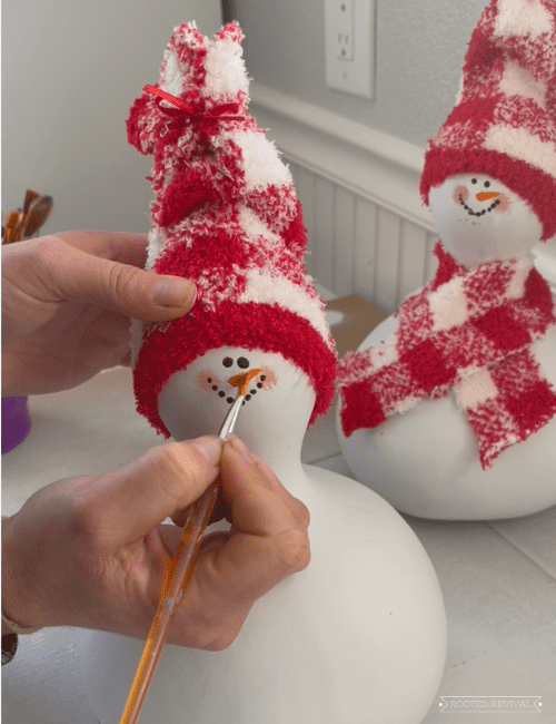 A woman holds a paint brush and paints a nose on a snowman gourd. There is a finished snowman gourd in the background.