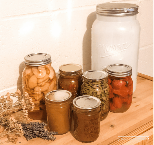 Several jars of canned food sit on a wooden shelf. There are dried flowers and a large white mason jar nearby.