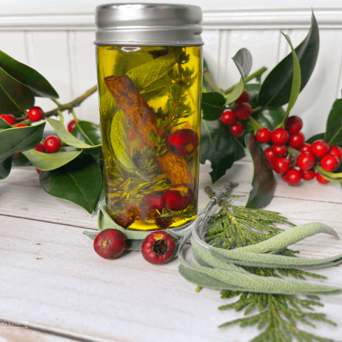 A small jar sits on a table adorned with fresh holly and berries. In the jar is yellow oil with a cinnamon stick, herbs and berries suspended in it.
