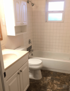 Behind the Scenes August 2019: Bathroom with white tile in the shower, white vanity and brown vinyl flooring