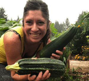 Kaylee holding up two large zucchini squash. She is smiling and wearing a yellow tank top.