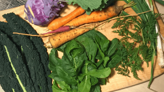 A wood cutting board loaded with dark leafy greens, kale, carrots, kohlrabi and green onions