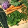 A wood cutting board loaded with dark leafy greens, kale, carrots, kohlrabi and green onions