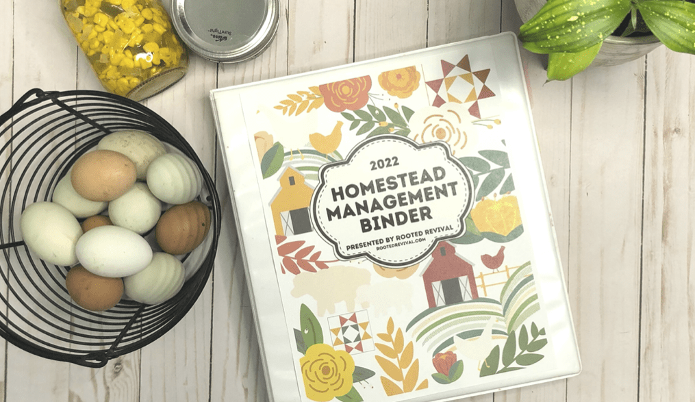 A white binder site on a pale wood background surrounded by an egg basket, a green plant and two glass jars of canned food. The cover of the binder shows images of farm scenes and has the title: 2022 Homestead Management Binder