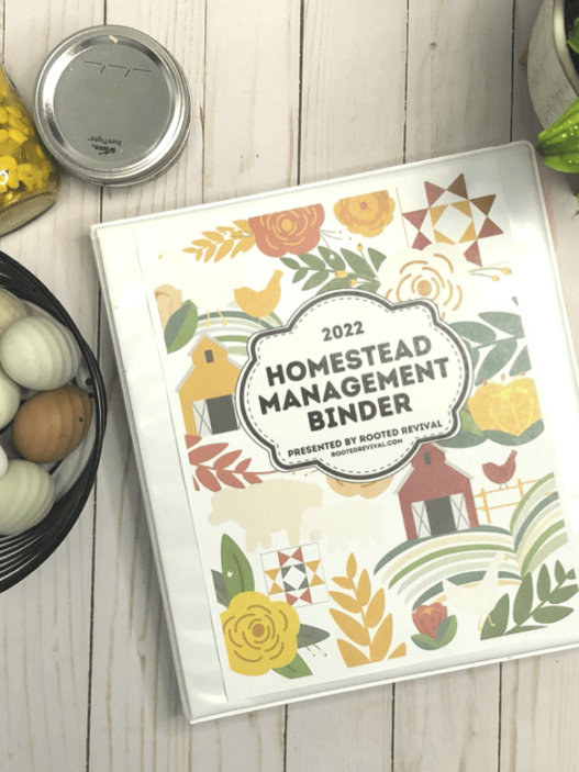 A white binder site on a pale wood background surrounded by an egg basket, a green plant and two glass jars of canned food. The cover of the binder shows images of farm scenes and has the title: 2022 Homestead Management Binder