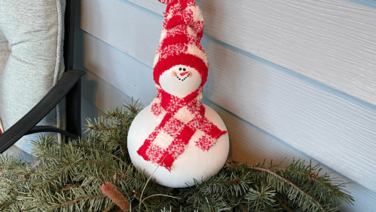 A snowman created from a gourd sits on a pine boughs. The snowman is painted white, has a painted face and red & white checkered scarf and hat made from a sock