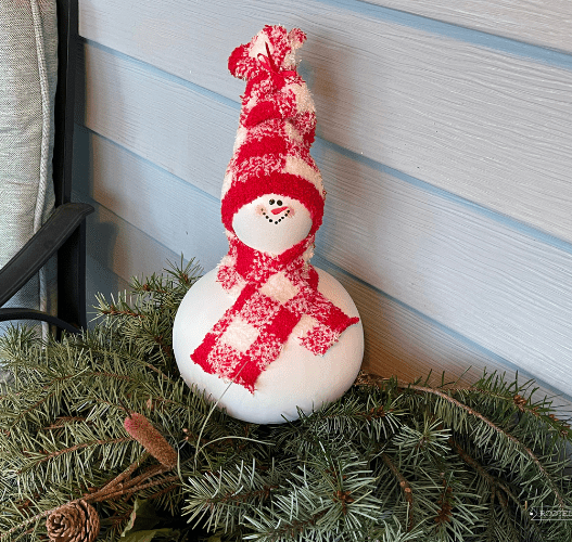 A snowman created from a gourd sits on a pine boughs. The snowman is painted white, has a painted face and red & white checkered scarf and hat made from a sock