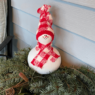 A snowman created from a gourd sits on a pine boughs. The snowman is painted white, has a painted face and red & white checkered scarf and hat made from a sock