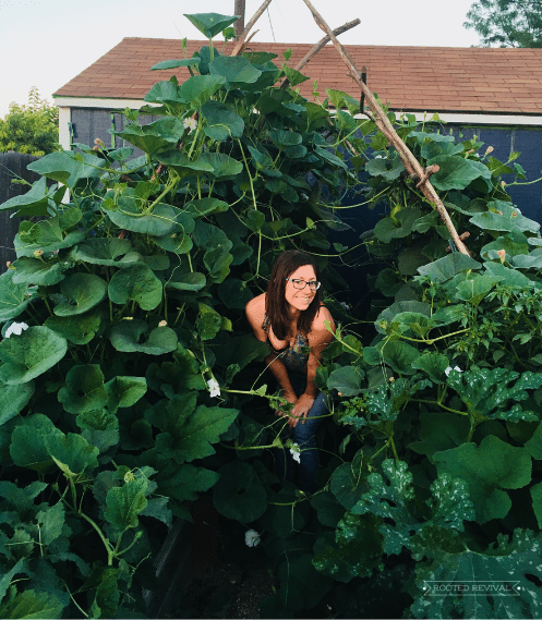 A woman with long brown hair wearing a colorful tank top crouches under an arched trellis filled with gourd vines