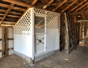 A small white chicken coop built inside a barn. The bottom half of the walls are solid wood and the top half are white lattice.