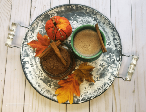 A shabby white round tray sits on white aged boards. The tray has a cup of hot chocolate and a jar with hot chocolate mix beside fall leaves and a small pumpkin