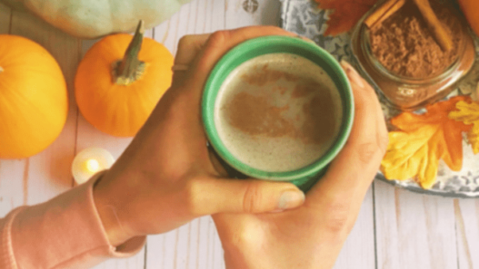 A set of woman's hands hold a cup of hot chocolate above some pumpkins and a glass jar of hot chocolate mix