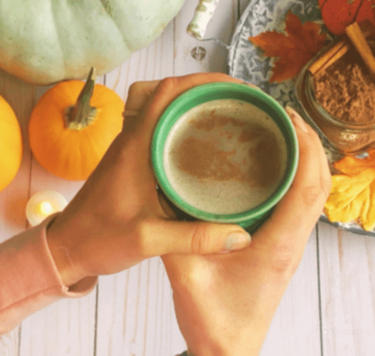 A set of woman's hands hold a cup of hot chocolate above some pumpkins and a glass jar of hot chocolate mix