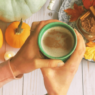 A set of woman's hands hold a cup of hot chocolate above some pumpkins and a glass jar of hot chocolate mix