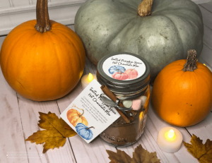 A glass jar sits in front of 2 orange and one blue pumpkin with tea lights around it. The glass jar has a tag that reads "Salted Pumpkin Spice Hot Chocolate Mix"