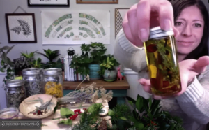 A screenshot shows Kaylee holding up a small jar of infused oil. In the background, there are lots of herbs and plants and jars of dried herbs arranged on a table.