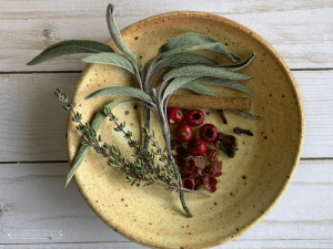 A small yellow hand-made pottery bowl holds some herb sprigs, a cinnamon stick, some berries and cloves