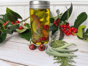 A small jar sits on a table adorned with fresh holly and berries. In the jar is yellow oil with a cinnamon stick, herbs and berries suspended in it.