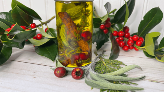 A small jar sits on a table adorned with fresh holly and berries. In the jar is yellow oil with a cinnamon stick, herbs and berries suspended in it.