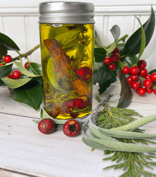 A small jar sits on a table adorned with fresh holly and berries. In the jar is yellow oil with a cinnamon stick, herbs and berries suspended in it.