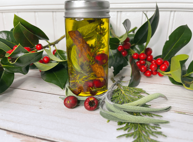 A small jar sits on a table adorned with fresh holly and berries. In the jar is yellow oil with a cinnamon stick, herbs and berries suspended in it.