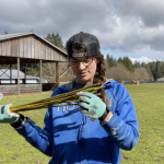 A woman (Lindy) hold up a handful of willow cuttings to inspect them. She is wearing a backwards baseball cap, a blue hoodie and blue work gloves