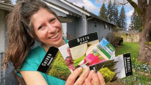 A woman (Kaylee) holds up an assortment of seed packets. She is standing outside in front of a blue house and is wearing a blue shirt.