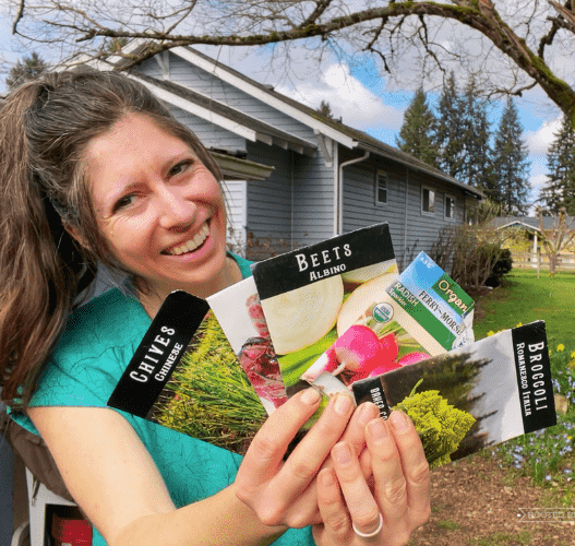 A woman (Kaylee) holds up an assortment of seed packets. She is standing outside in front of a blue house and is wearing a blue shirt.