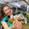 A woman (Kaylee) holds up an assortment of seed packets. She is standing outside in front of a blue house and is wearing a blue shirt.