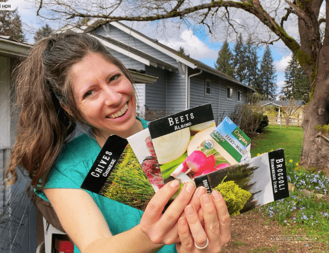 A woman (Kaylee) holds up an assortment of seed packets. She is standing outside in front of a blue house and is wearing a blue shirt.
