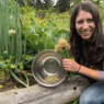 Kaylee sits in the garden by a garden bed. She holds 3 small green onion blossoms in one hand and a steel bowl in her other hand. The bowl has some seeds and plant matter in it.