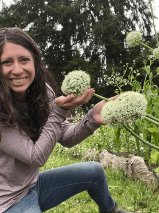 Kaylee sits in the garden and cups giant round onion blossoms in her hands. She is wearing a light pink shirt and blue jeans.