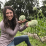 Kaylee sits in the garden and cups giant round onion blossoms in her hands. She is wearing a light pink shirt and blue jeans.